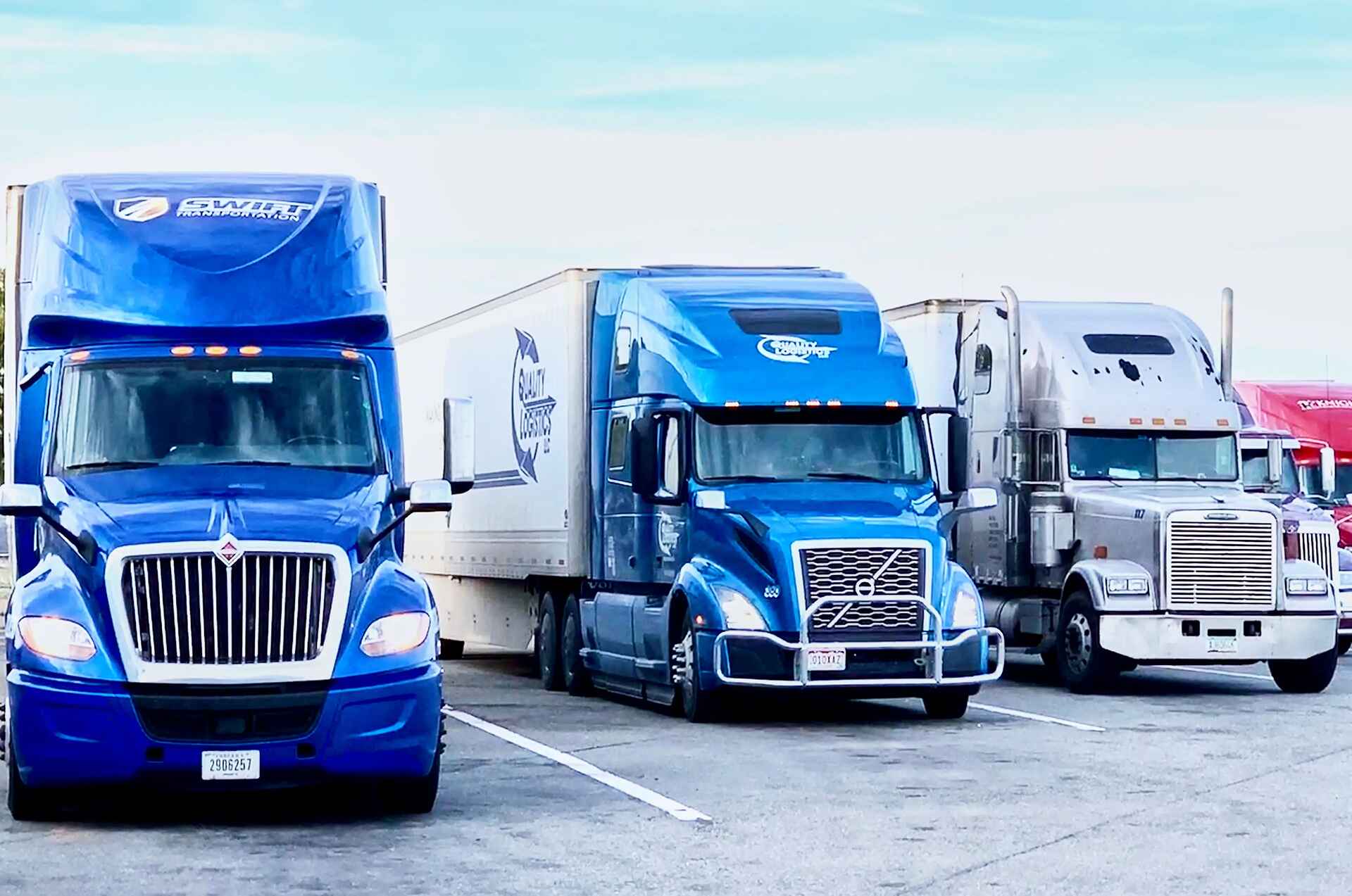 two blue semi trucks, a silver semi truck and a red semi truck parked in a parking lot